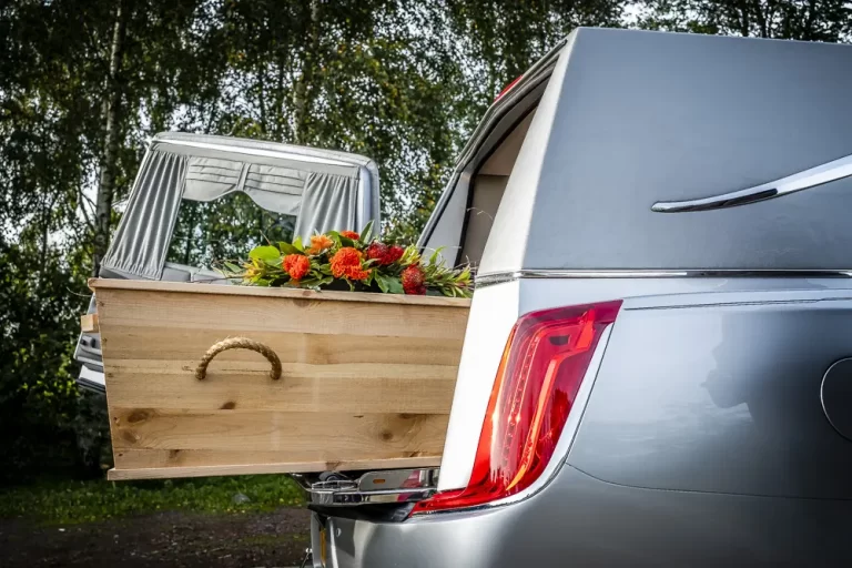 Een houtengrafkist in een lichtgrijze rouwauto, uitvaart in scene gezet
